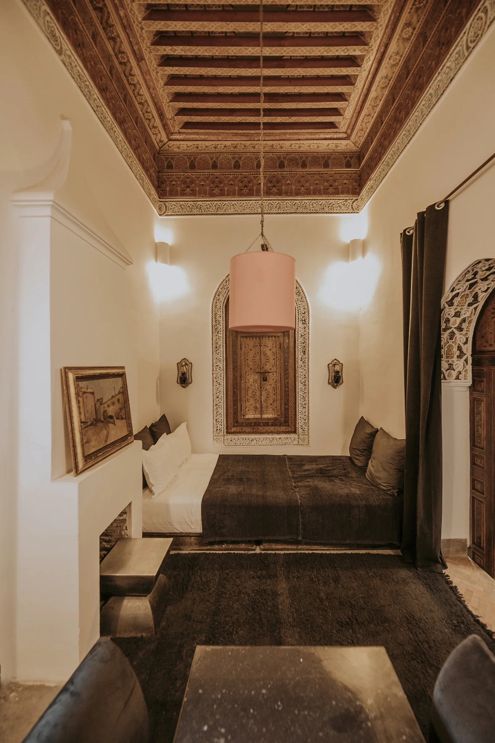 A room of Dar Zelda beneath its painted-cedar ceiling — dark silks and wools over the bed, a fireplace beside it
